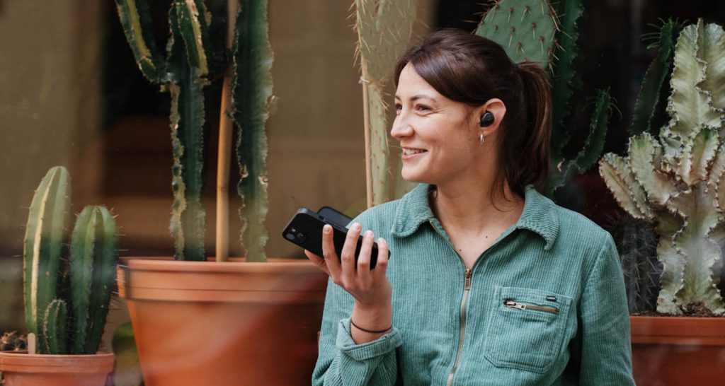 Una mujer sonriente lleva auriculares in-ear y un smartphone en la mano.