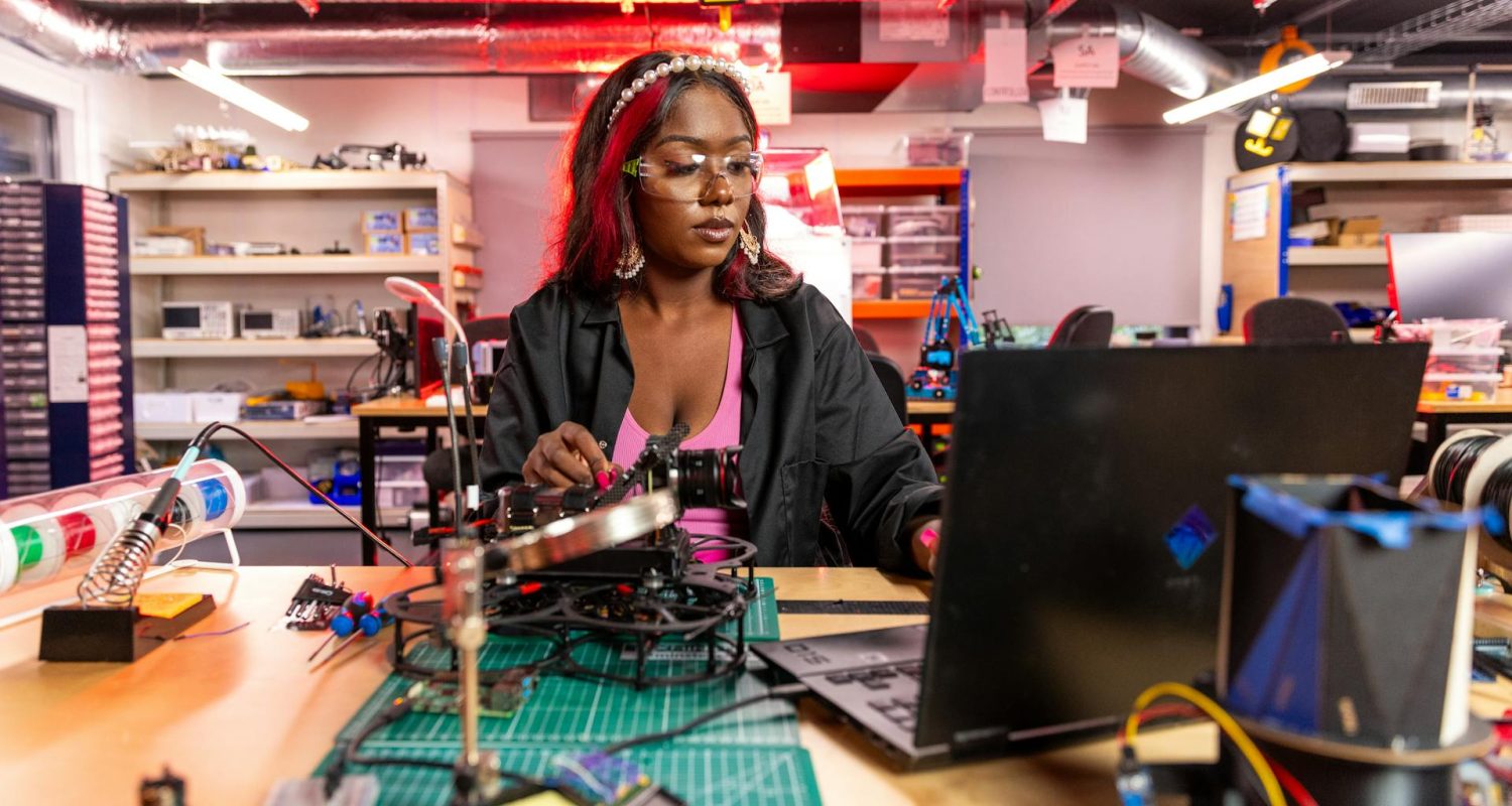 Frau arbeitet in einer Werkstatt an technischen Bauteilen und einem Laptop