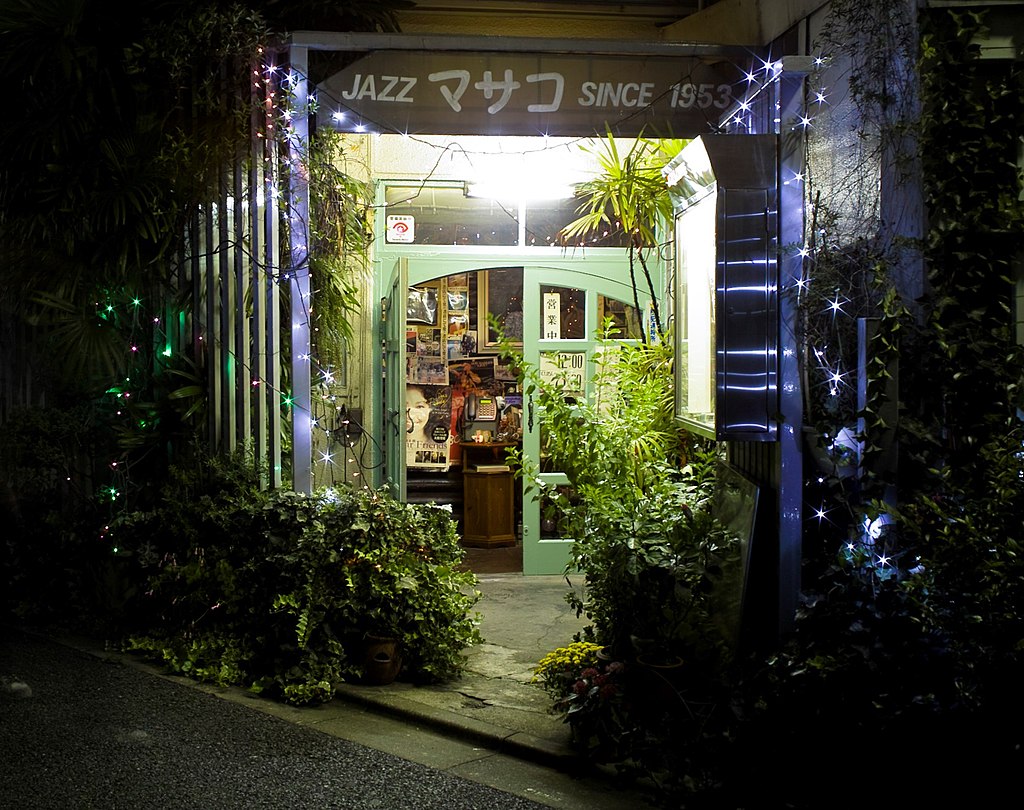 Entrada a un café jazz con iluminación ambiental, puerta verde, plantas y guirnaldas luminosas.