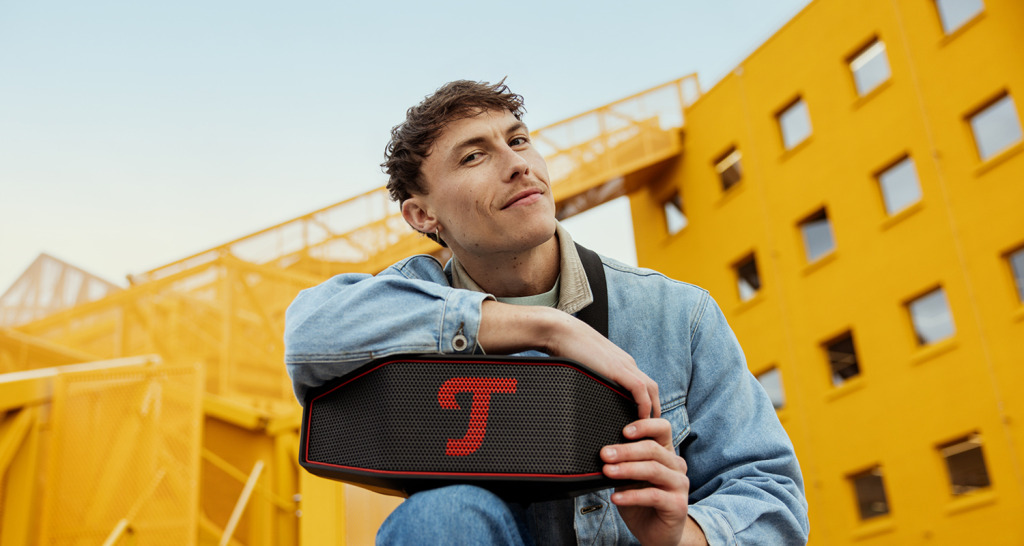 ROCKSTER CROSS 2 Un hombre posa con el ROCKSTER CROSS 2 de Teufel delante de un edificio amarillo.